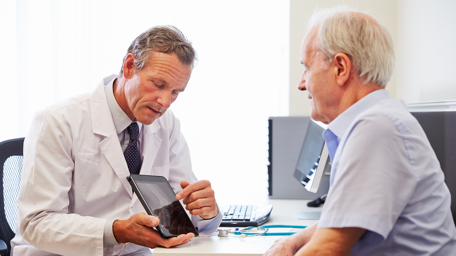 Læge, der viser noget på en tablet til en ældre herre inde på sit kontor