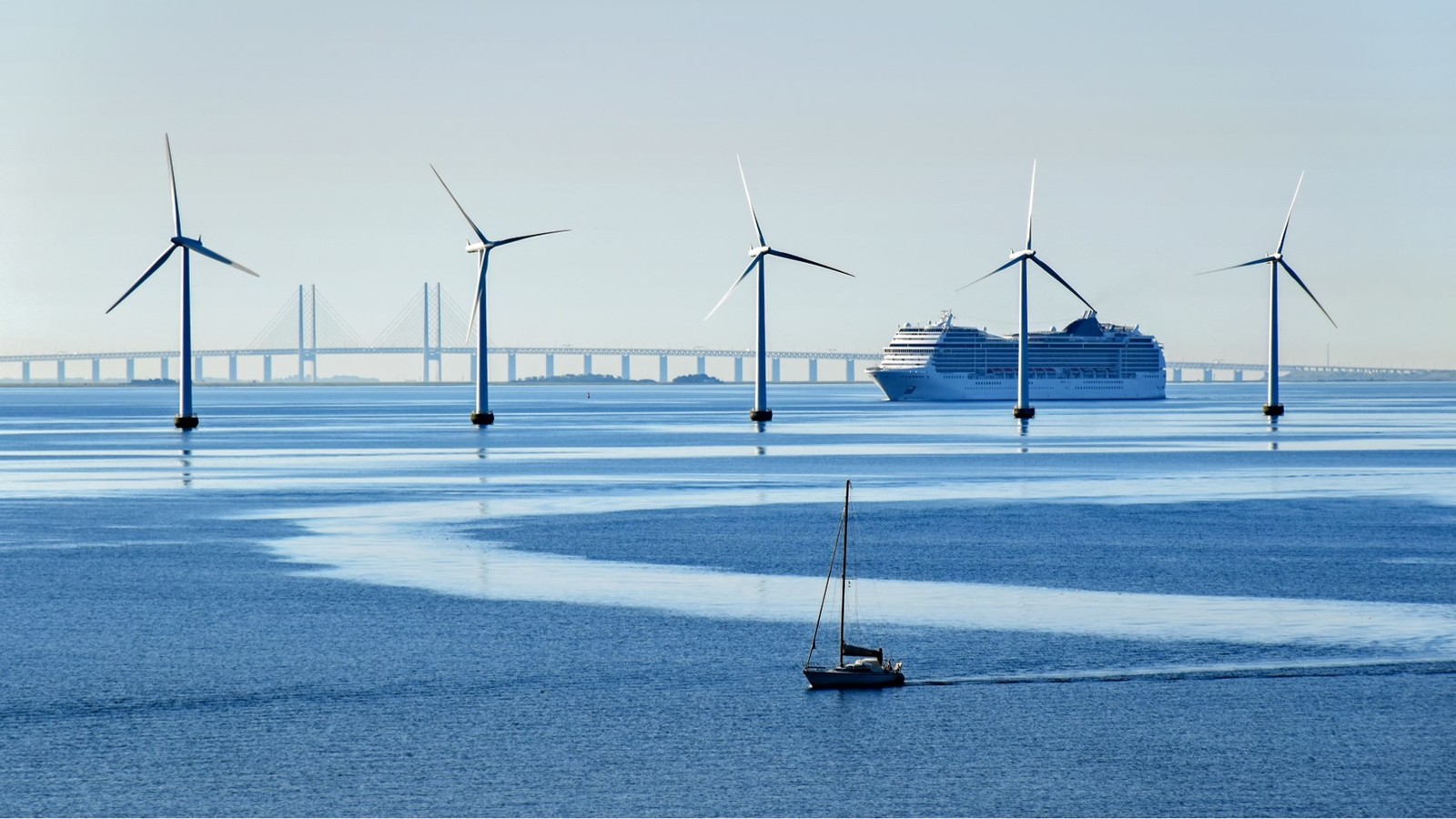 Fem vindmøller på linje i vandet med sejlbåd i forgrunden samt færge og Storebæltsbroen i baggrunden