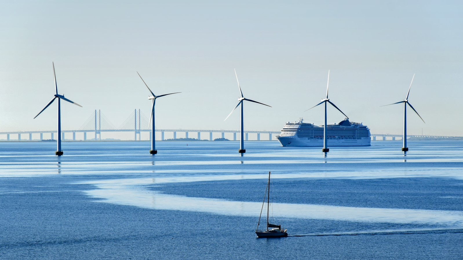 5 vindmøller i vandet med en sejlbåd i forgrunden samt en færge og Storebæltsbroen i baggrunden