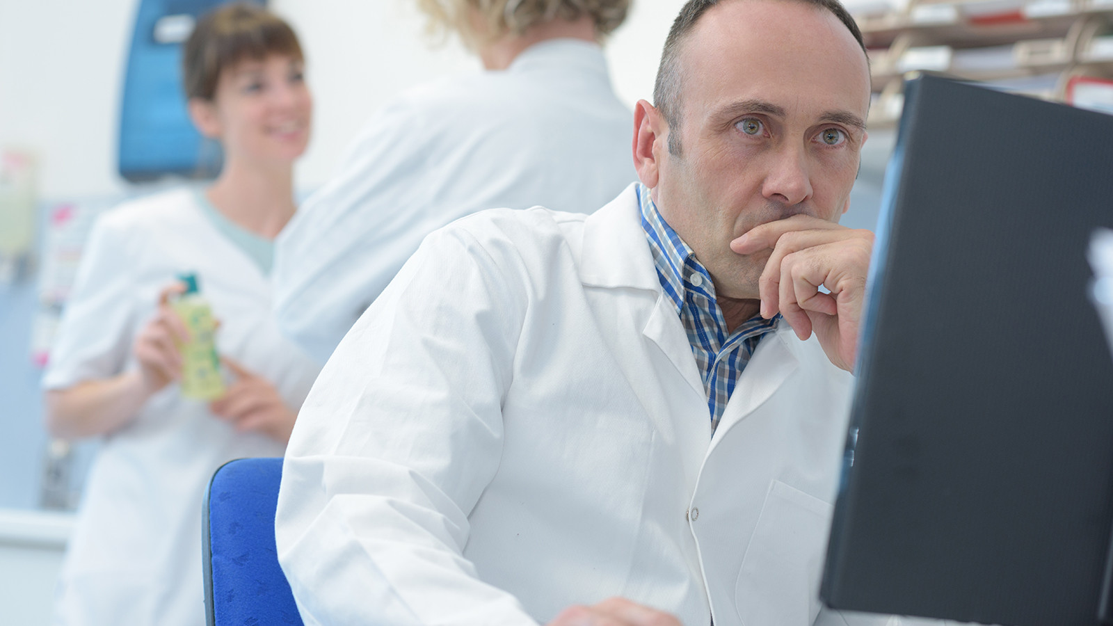Mandlig læge kigger koncentreret på sin computerskærm med kvindelige kollegaer, der taler sammen, i baggrunden